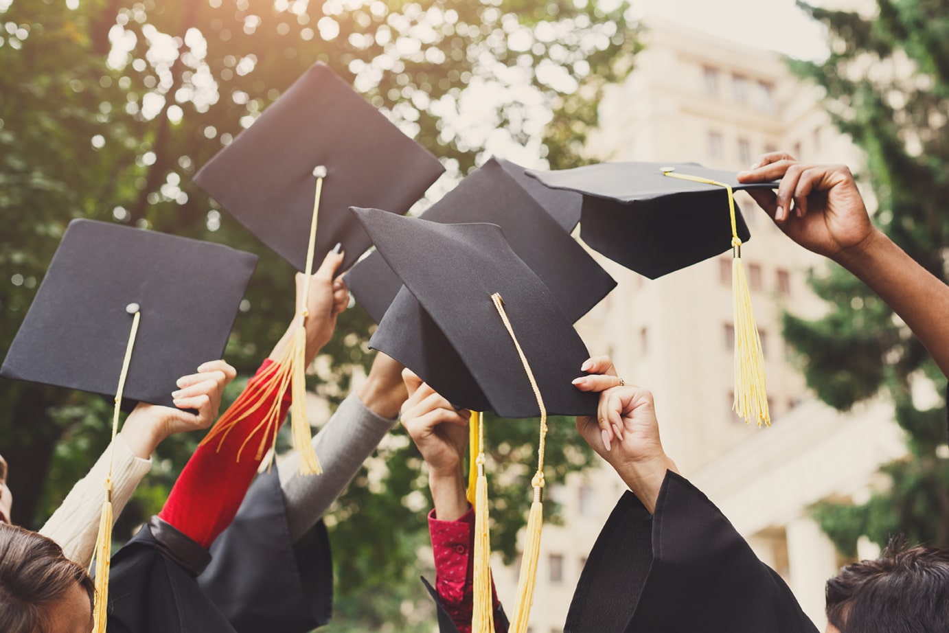 Group of graduates raising their black mortarboards with gold tassels in celebration, with trees and a university building in the background.