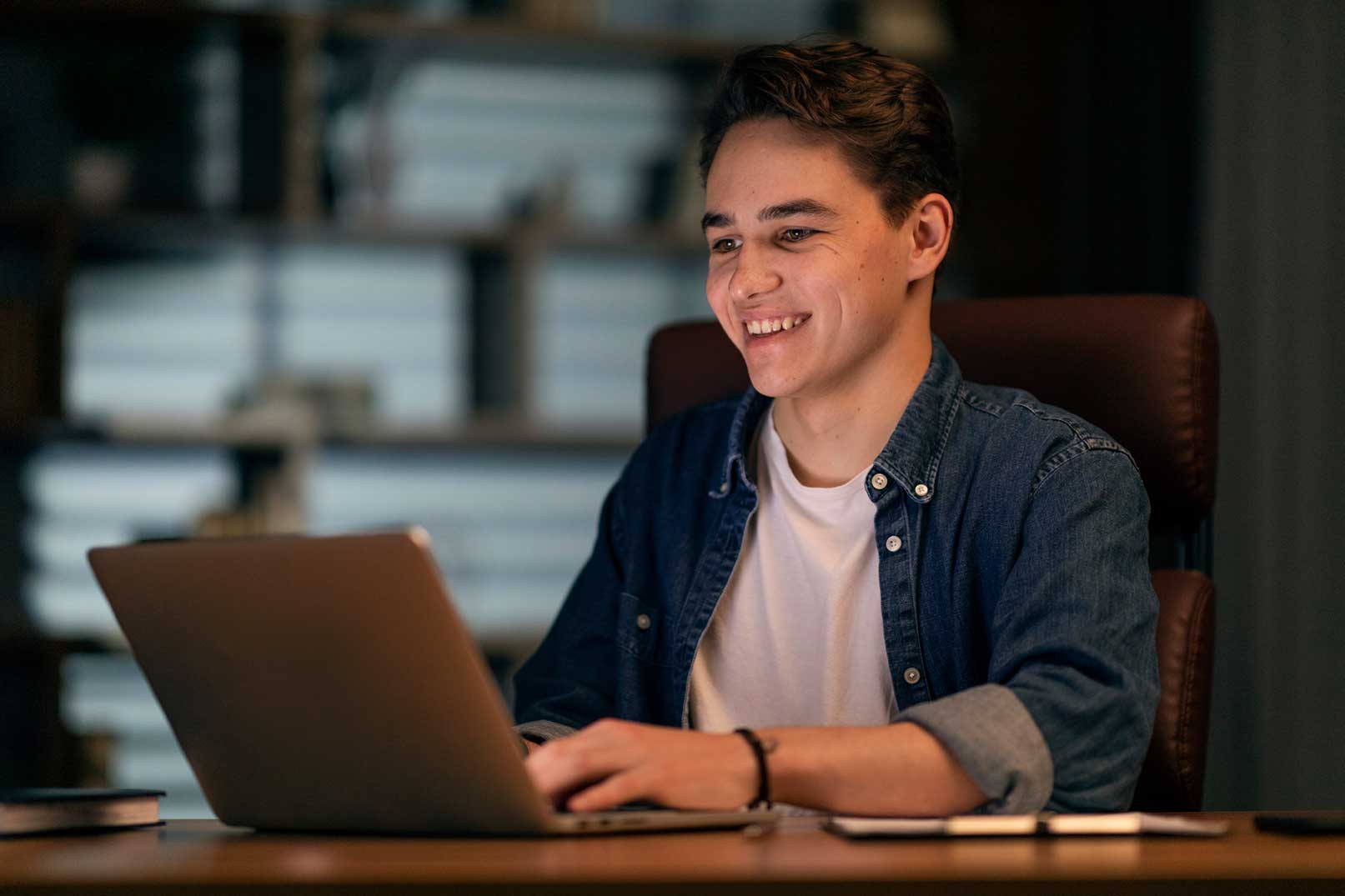 Smiling young man using a laptop at a desk in a dimly lit room with bookshelves in the background.