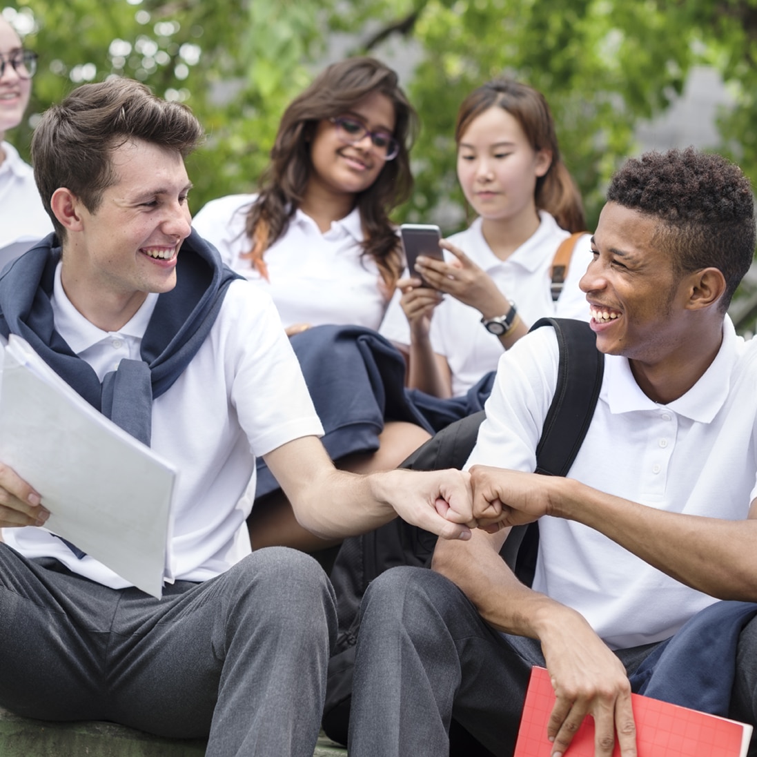 Group of students in school uniforms sitting outdoors, with two male students smiling and fist-bumping in the foreground, and others engaging with phones and books in the background.