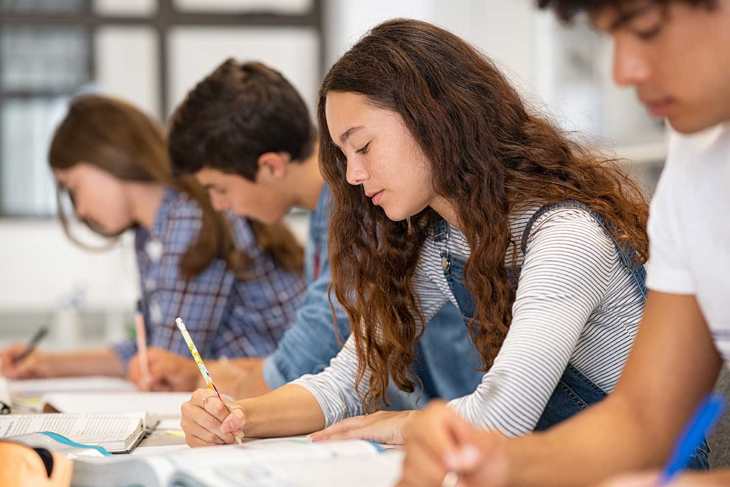 Group of high school students sitting at desks in a classroom, writing in notebooks and focusing on their work.