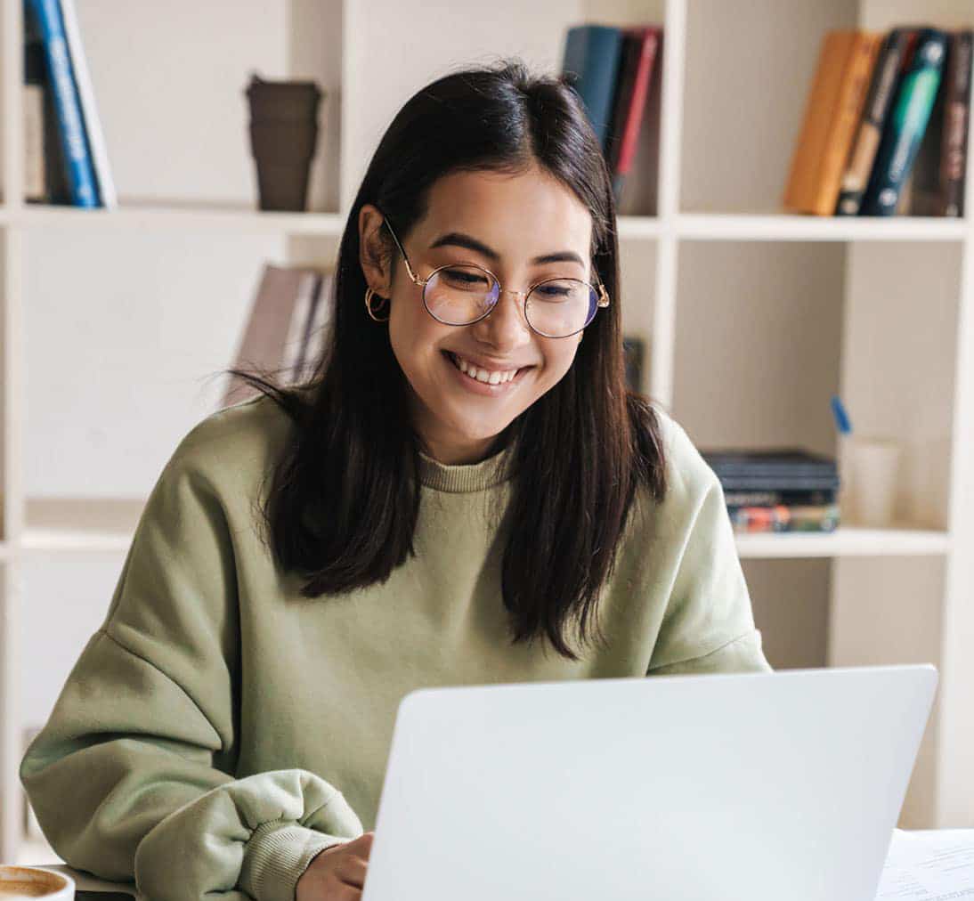 Happy young woman studying on laptop in library with coffee and books.